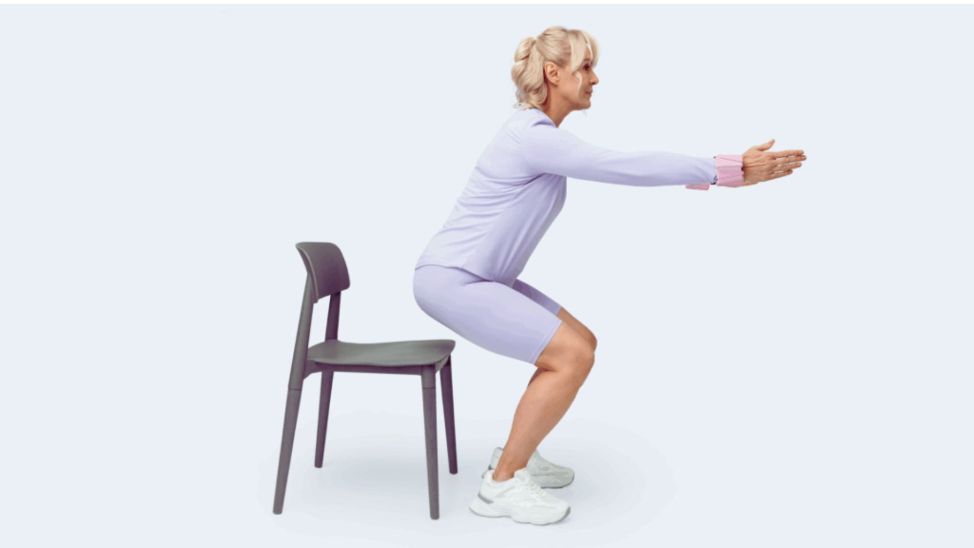 A woman performs a squat exercise using a chair for support in a fitness setting.
