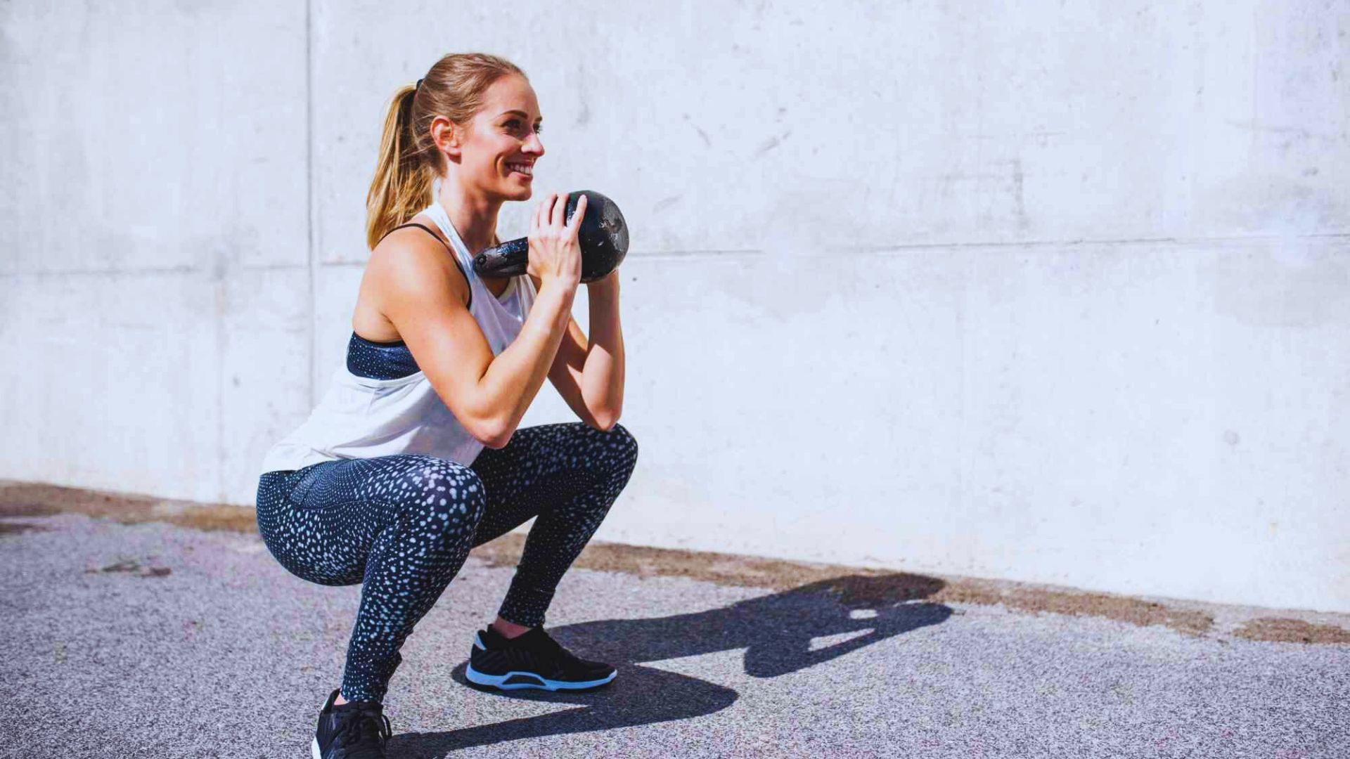 A woman performs a goblet squat while holding a kettlebell close to her chest in a gym setting.