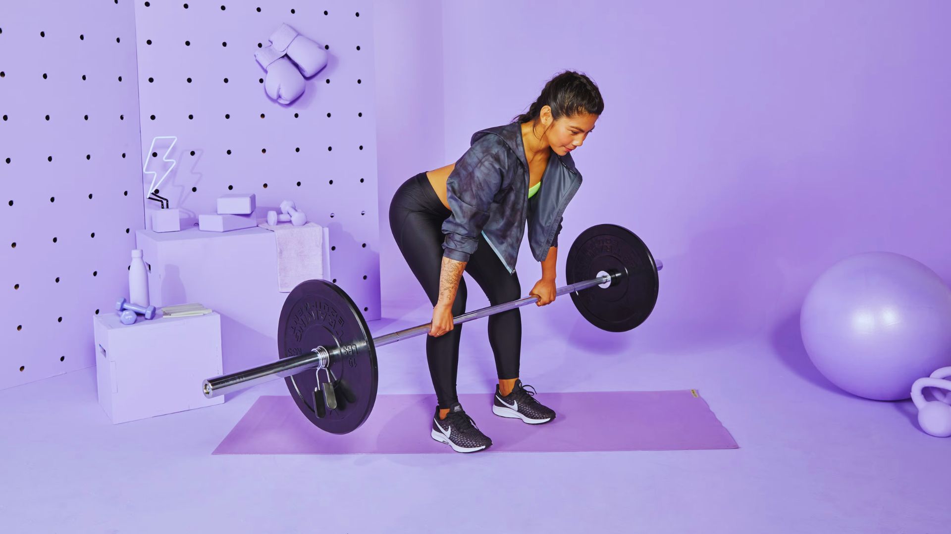 A girl is performing a deadlift with a barbell on a purple mat in a room decorated with purple polka dots and a lightning bolt neon sign.