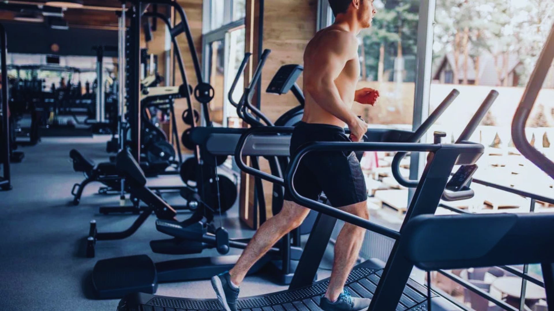 A man is jogging on a treadmill in a modern gym, focused on his workout amidst various gym equipment.
