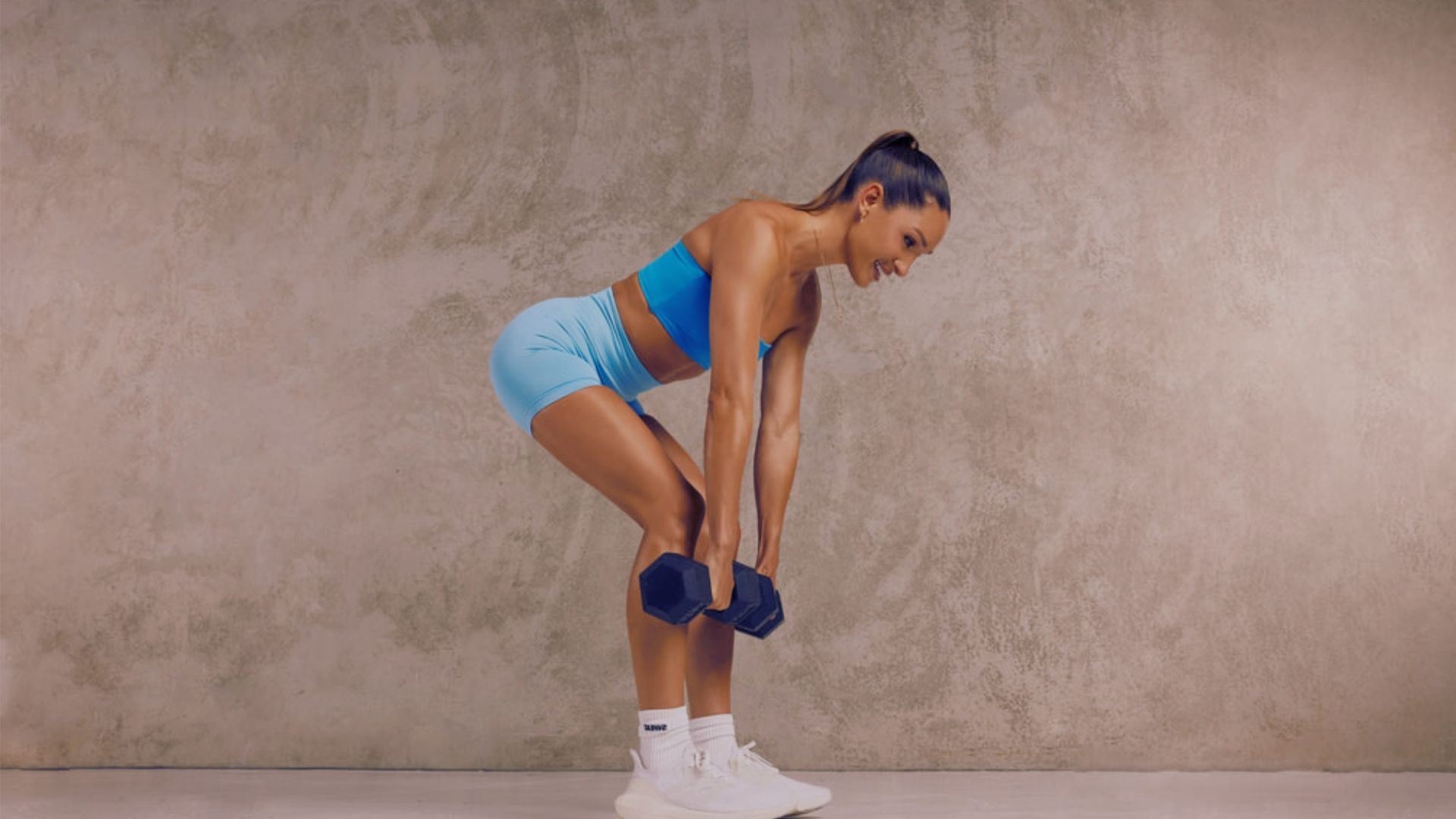 A woman in blue shorts and a blue top performs a squat, focusing on her form and balance in a fitness setting.