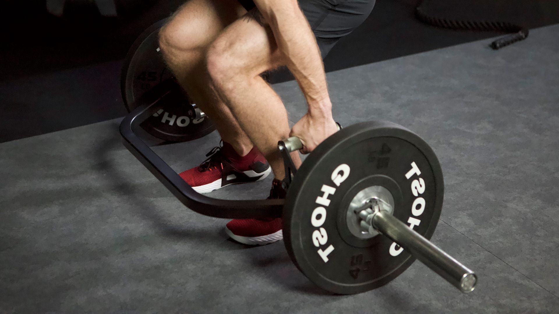 A man squats down while gripping a trap bar, demonstrating proper form for deadlifts in a gym setting.
