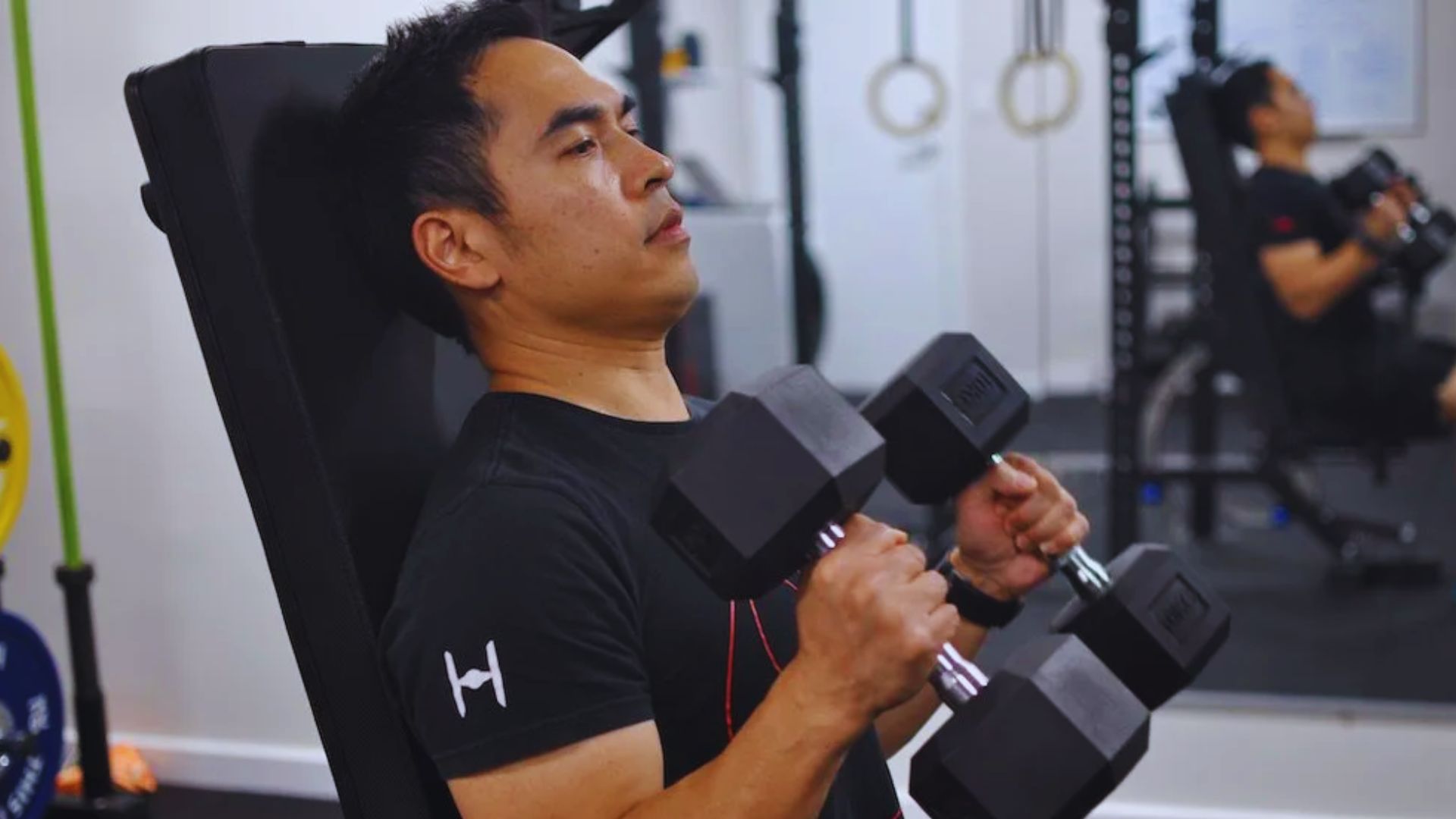 In a gym, a man uses a dumbbell while ensuring proper alignment by adjusting the seat and arm pads.