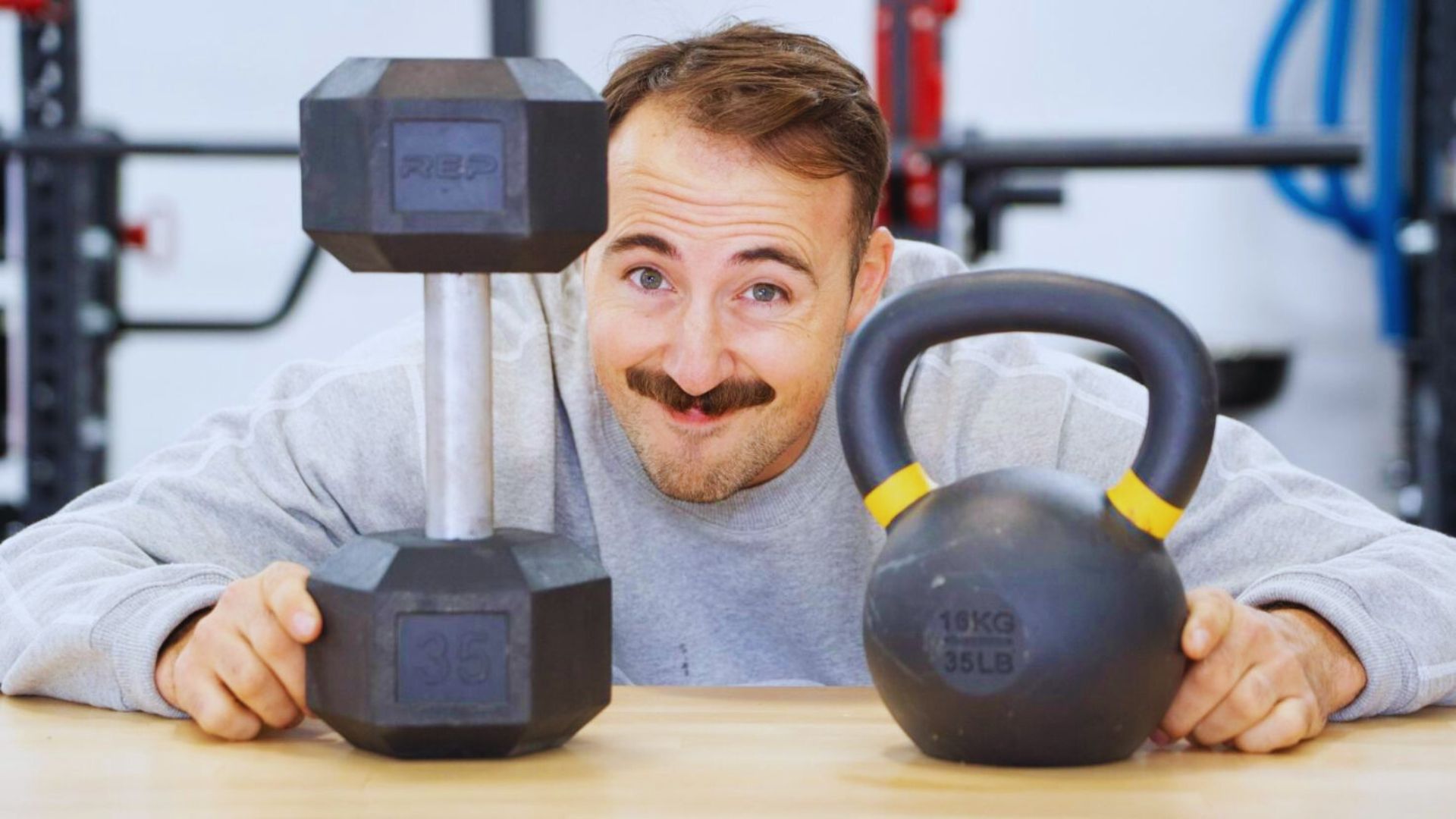 A man with a mustache holds two kettlebells, demonstrating strength and fitness in a gym setting.
