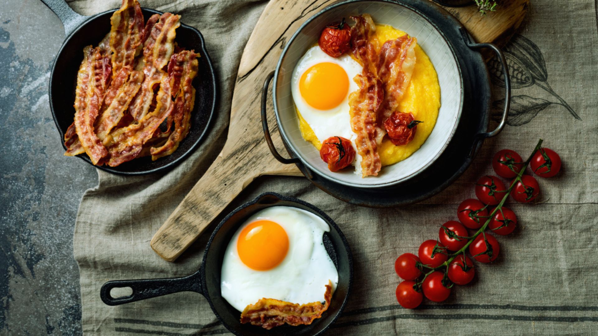 Fried eggs, crispy bacon, and ripe tomatoes served in cast iron skillets on a rustic wooden table.