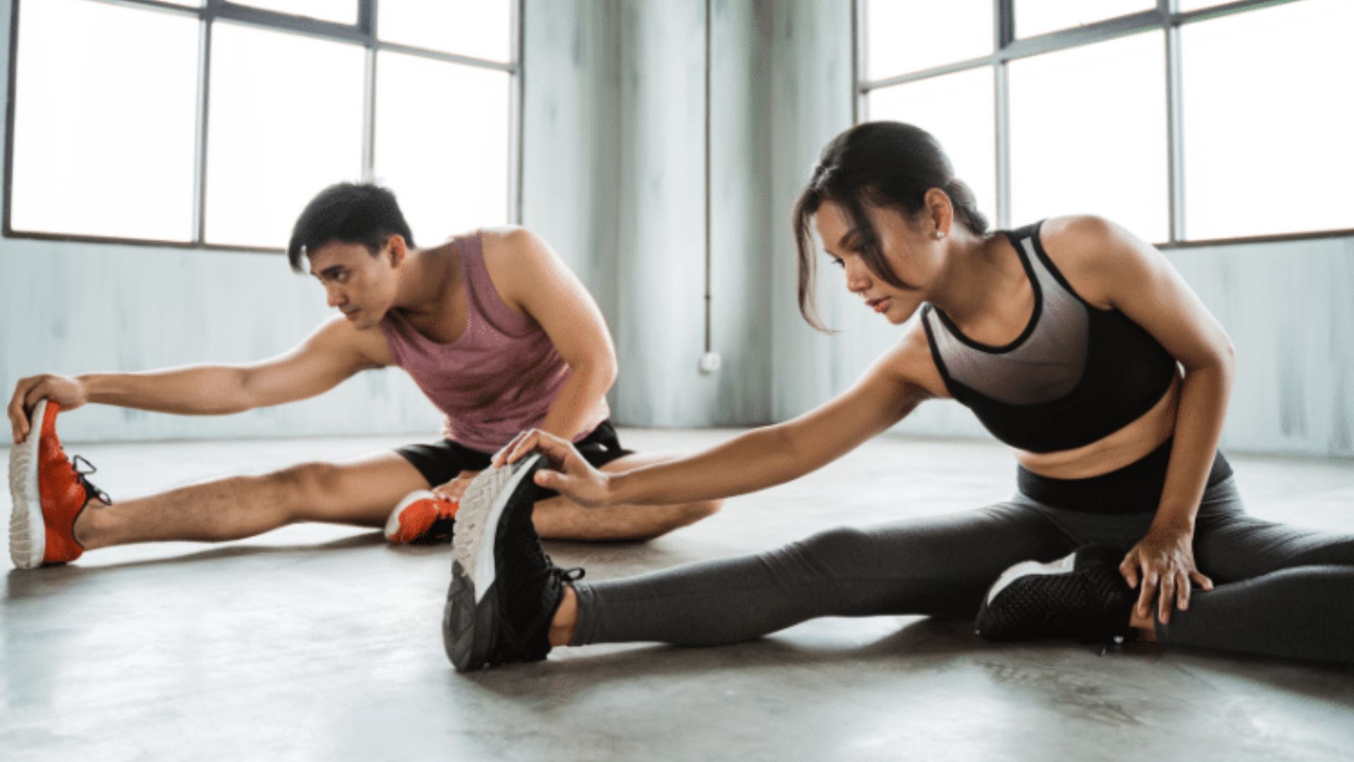 A man and woman perform stretching exercises together in a gym setting, focusing on flexibility and fitness.