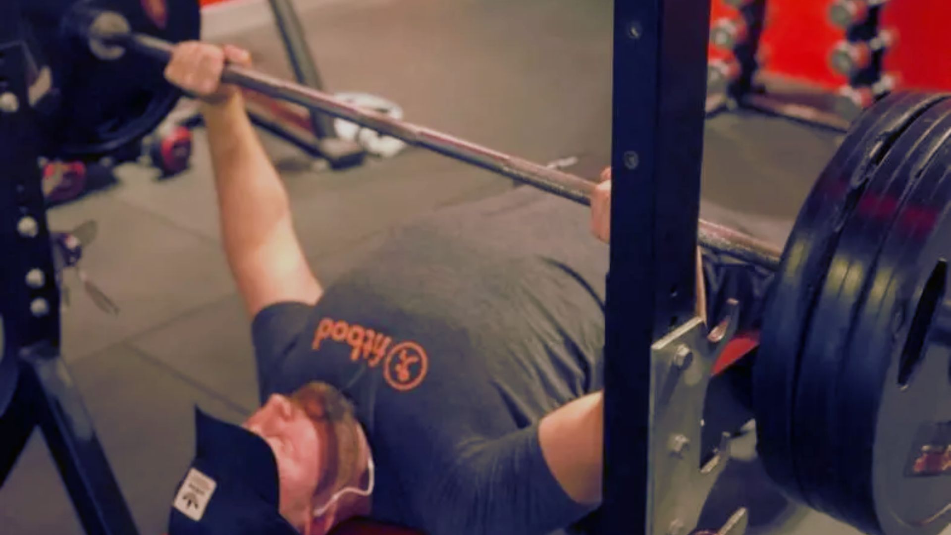 A man performs a bench press in a gym, focusing on strength training with weights on the barbell.