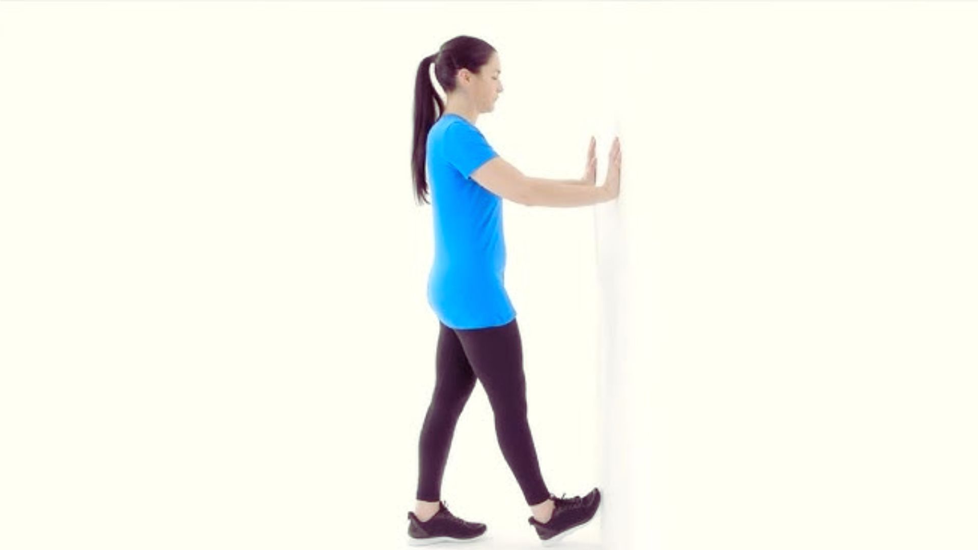 A woman stands against a wall, resting her hands on it, with a neutral expression on her face.