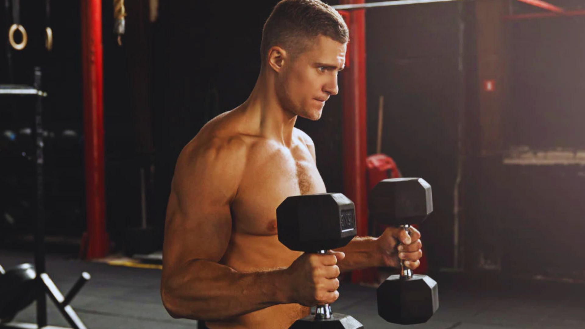 In a gym, a shirtless man holds two dumbbells for a hammer curl, showcasing proper grip and wrist alignment.