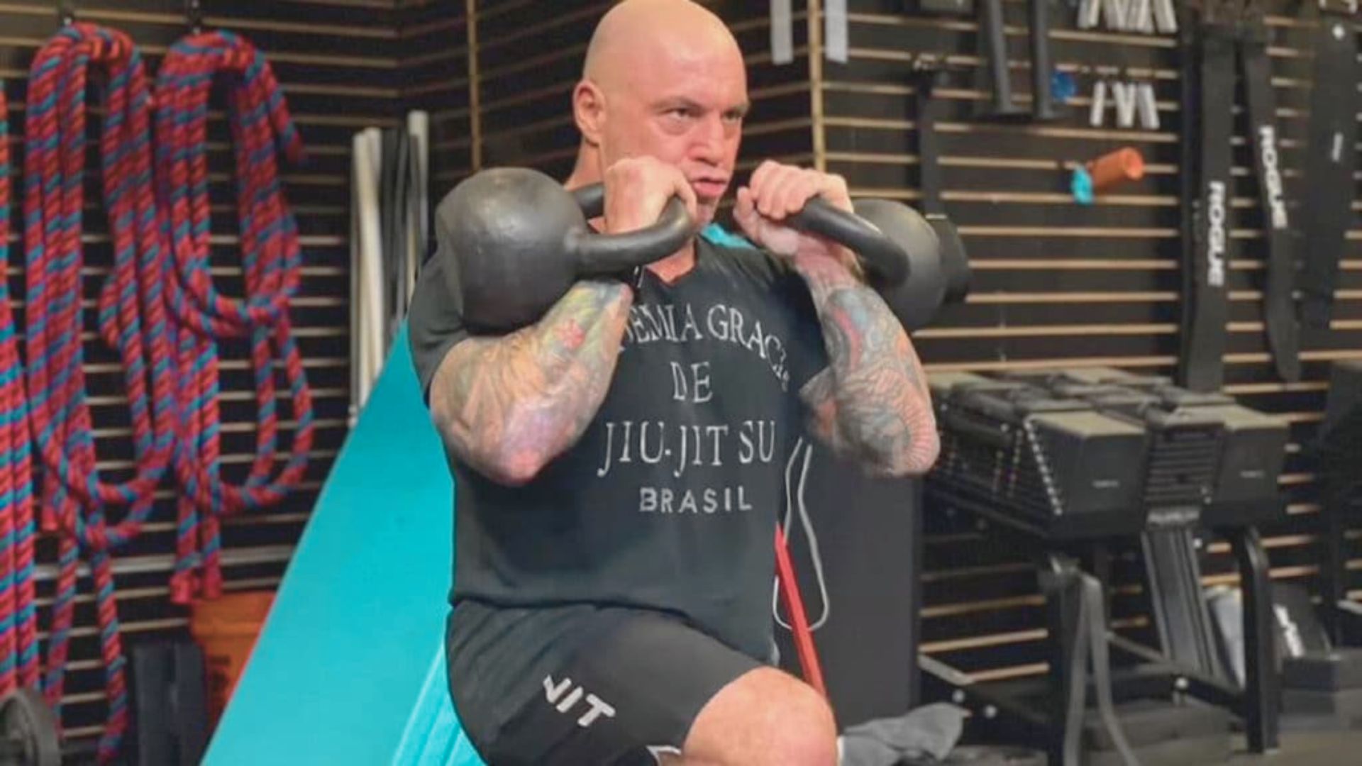A tattooed man performs a kettlebell exercise, focusing on strength training in a gym setting.