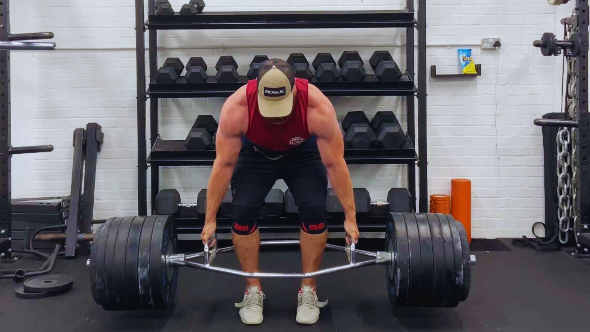 In a gym, a man is executing a trap bar squat while lifting a heavy bar.