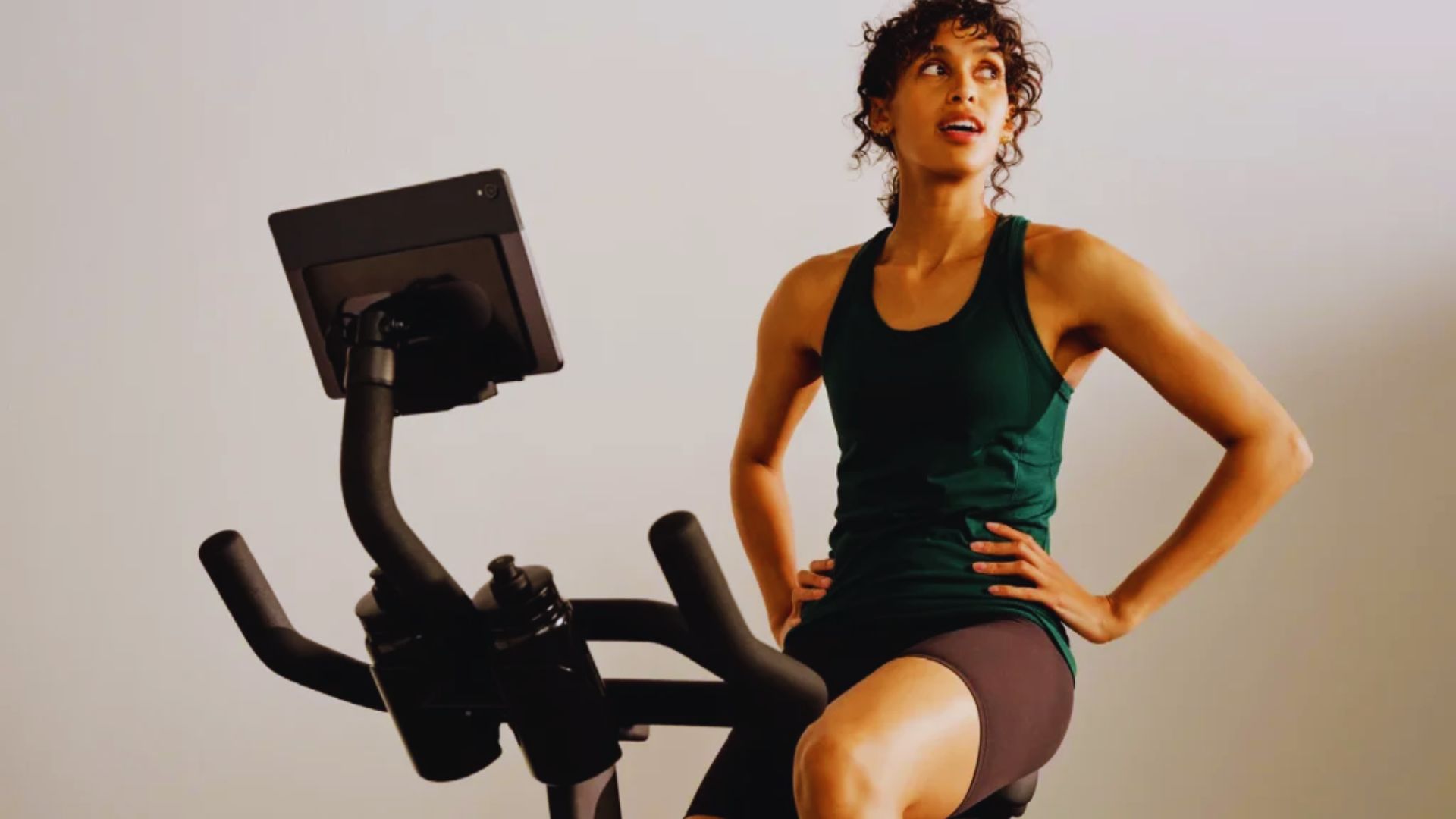 A woman sits on an exercise bike, focused on a tablet in her hands, engaged in a workout session.