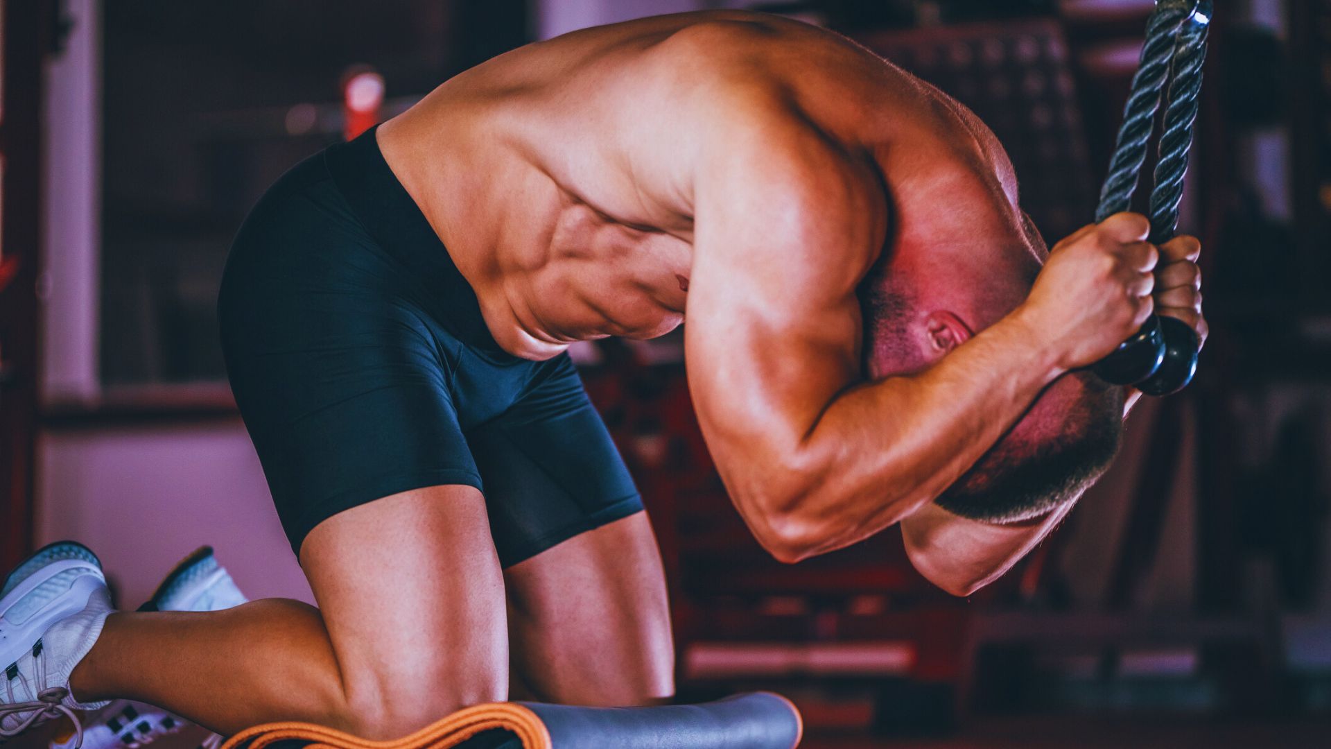 A man is kneeling on a gym bench, focused on his ab workout routine, with various exercise machines in the background.