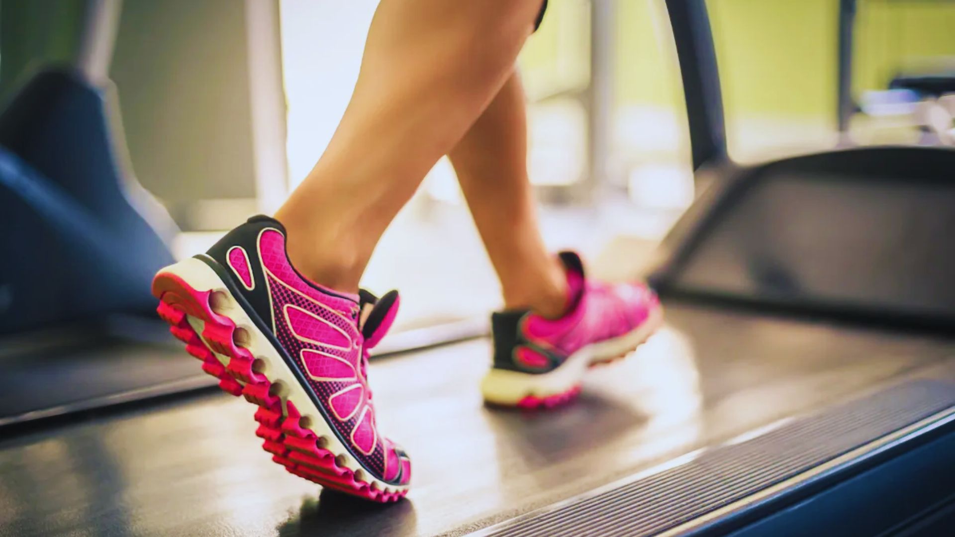 A woman in pink shoes exercises on a treadmill, focusing on her workout in a bright gym environment.