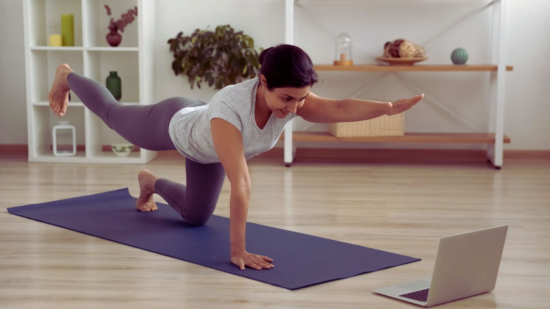 A woman practicing yoga on a mat while using a laptop for guidance or instruction.