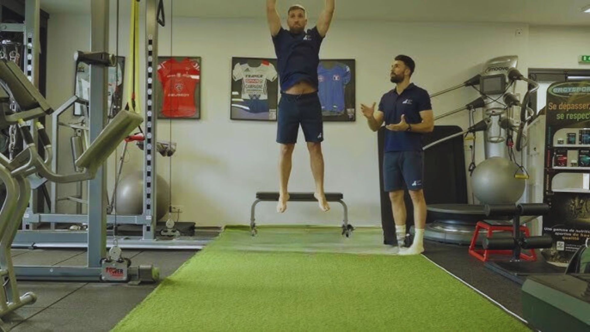 Two men exercising in a gym, one lifting weights while the other performs a stretching routine.