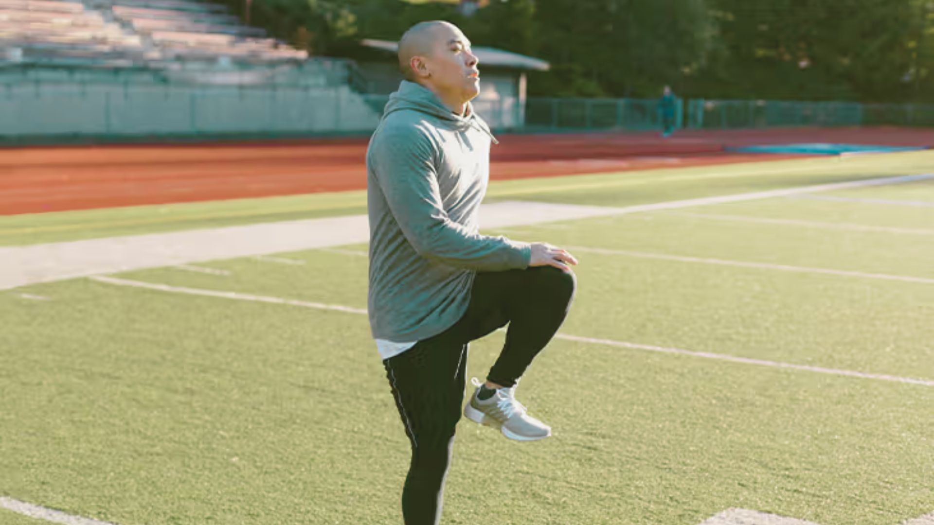 A man stretches on a football field, preparing for practice under a clear blue sky.