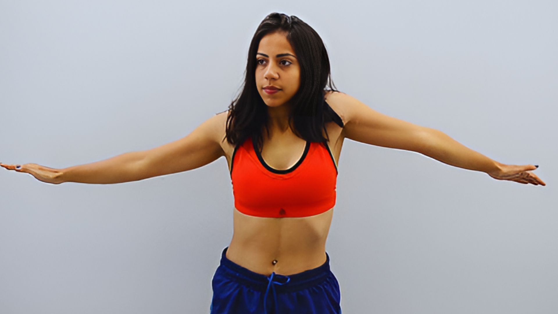 A woman wearing a sports bra and shorts engages in an arm movement exercise, showcasing her athleticism.