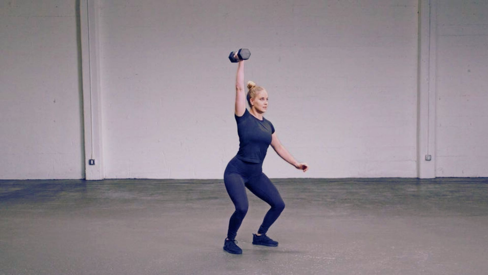A woman performs a DB power clean exercise in an empty room, focusing on her form and technique.