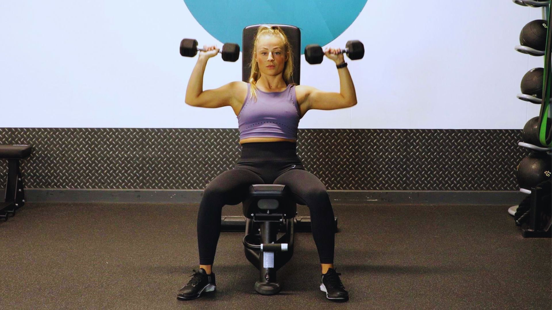 In a gym, a woman executes a dumbbell shoulder press, ensuring her seat height is adjusted for optimal performance.