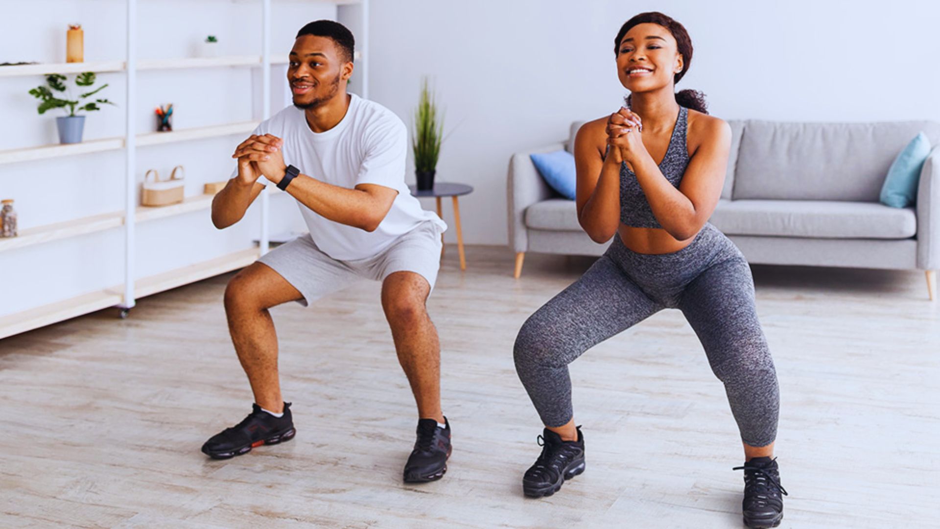 A man and woman performing squats together in a bright living room, surrounded by furniture and natural light.