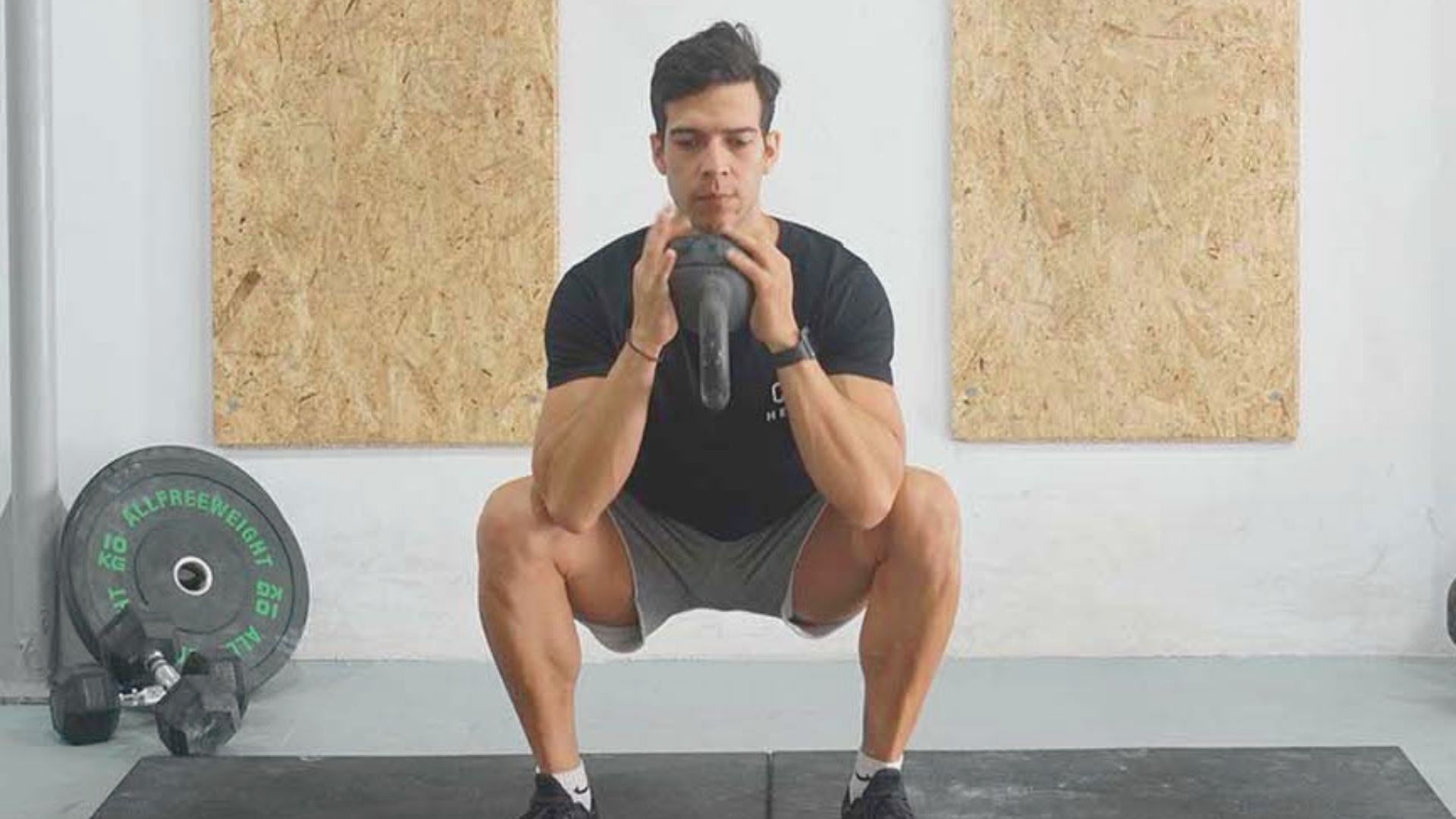 A man squats on a mat, holding a kettlebell in both hands, focusing on his workout routine.