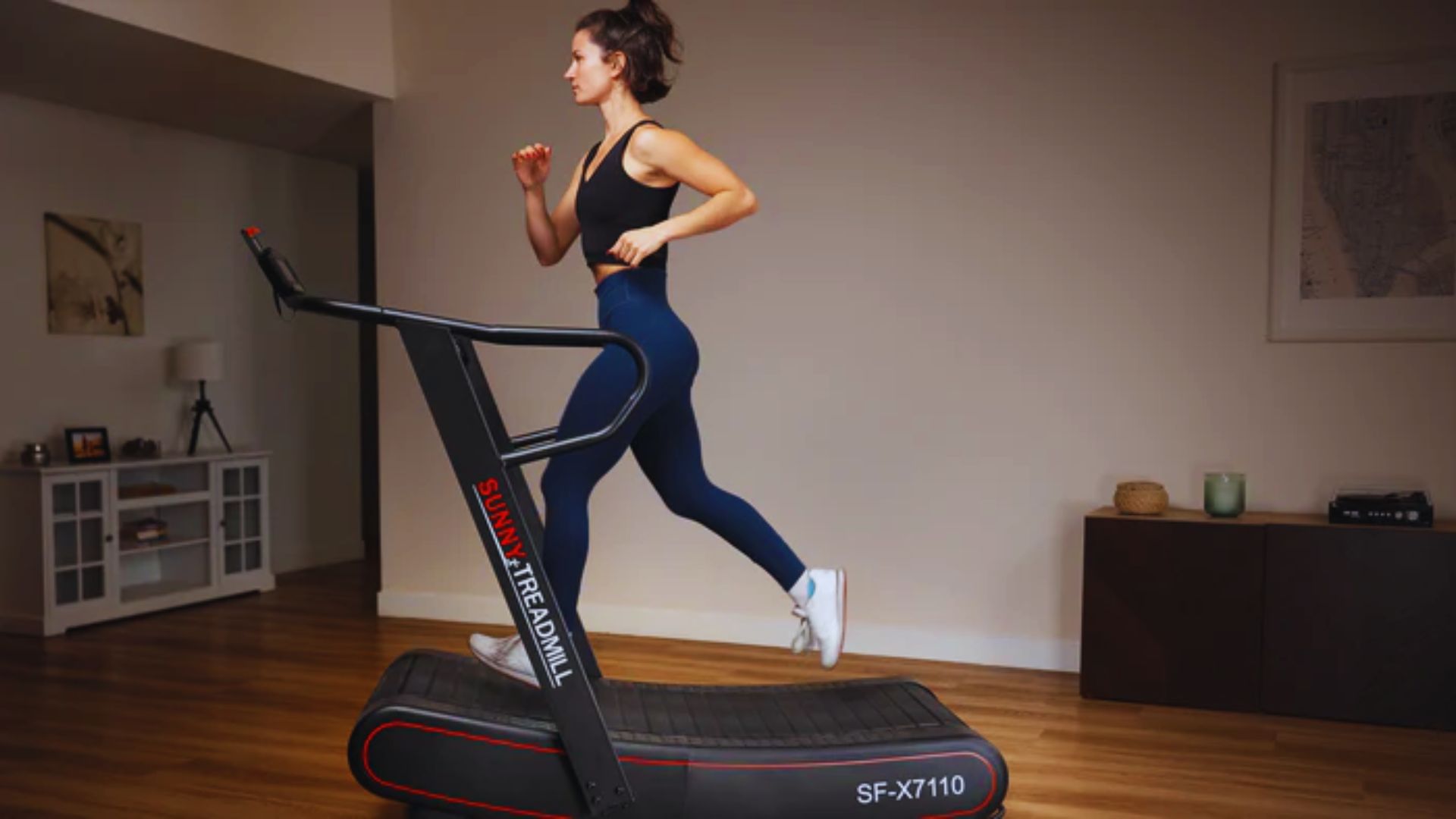 A woman runs on a treadmill in a cozy living room, surrounded by furniture and natural light.
