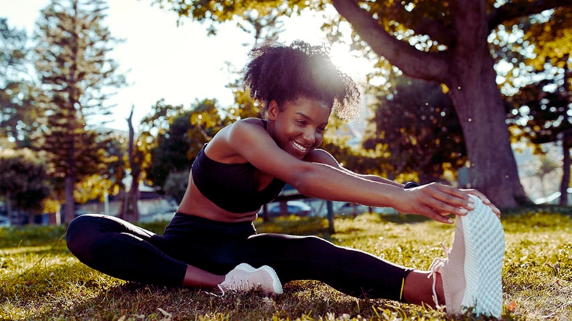 A woman stretches her arms and legs while sitting on the grass in a sunny outdoor setting