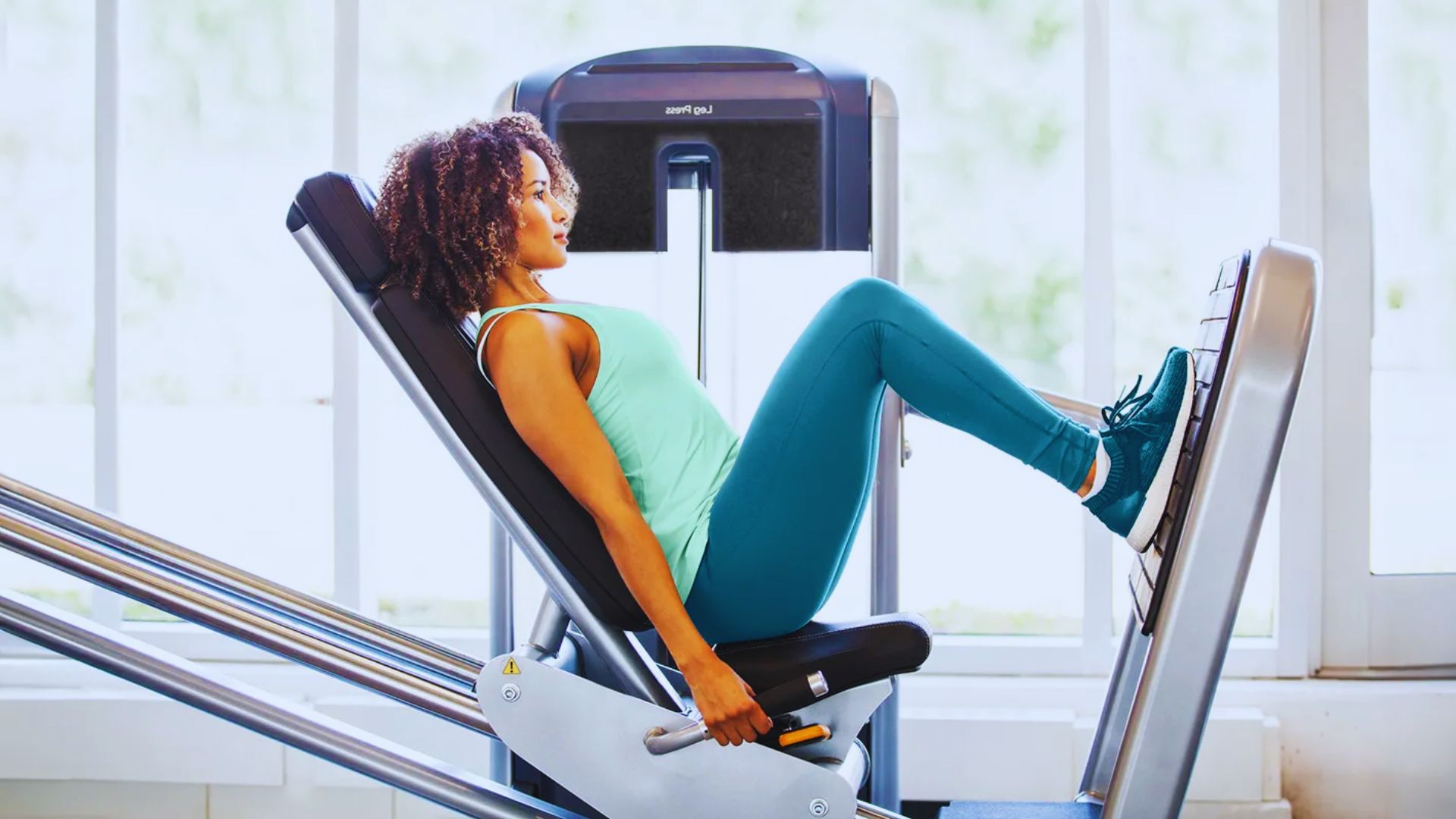 A woman sits on a gym machine, focusing on her workout while avoiding locking her knees at the top of the movement.