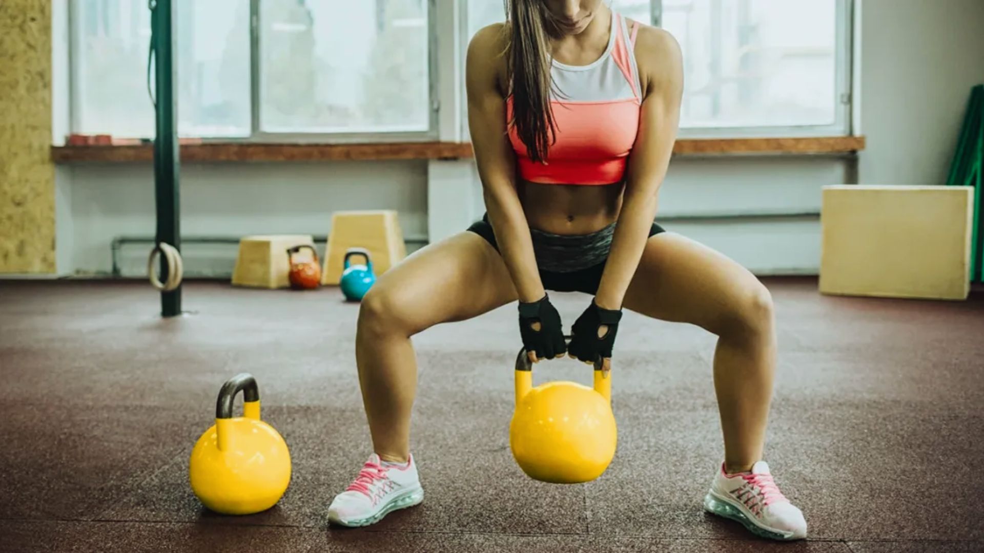 A woman squats while holding a kettlebell in each hand, demonstrating strength and fitness in a gym setting.