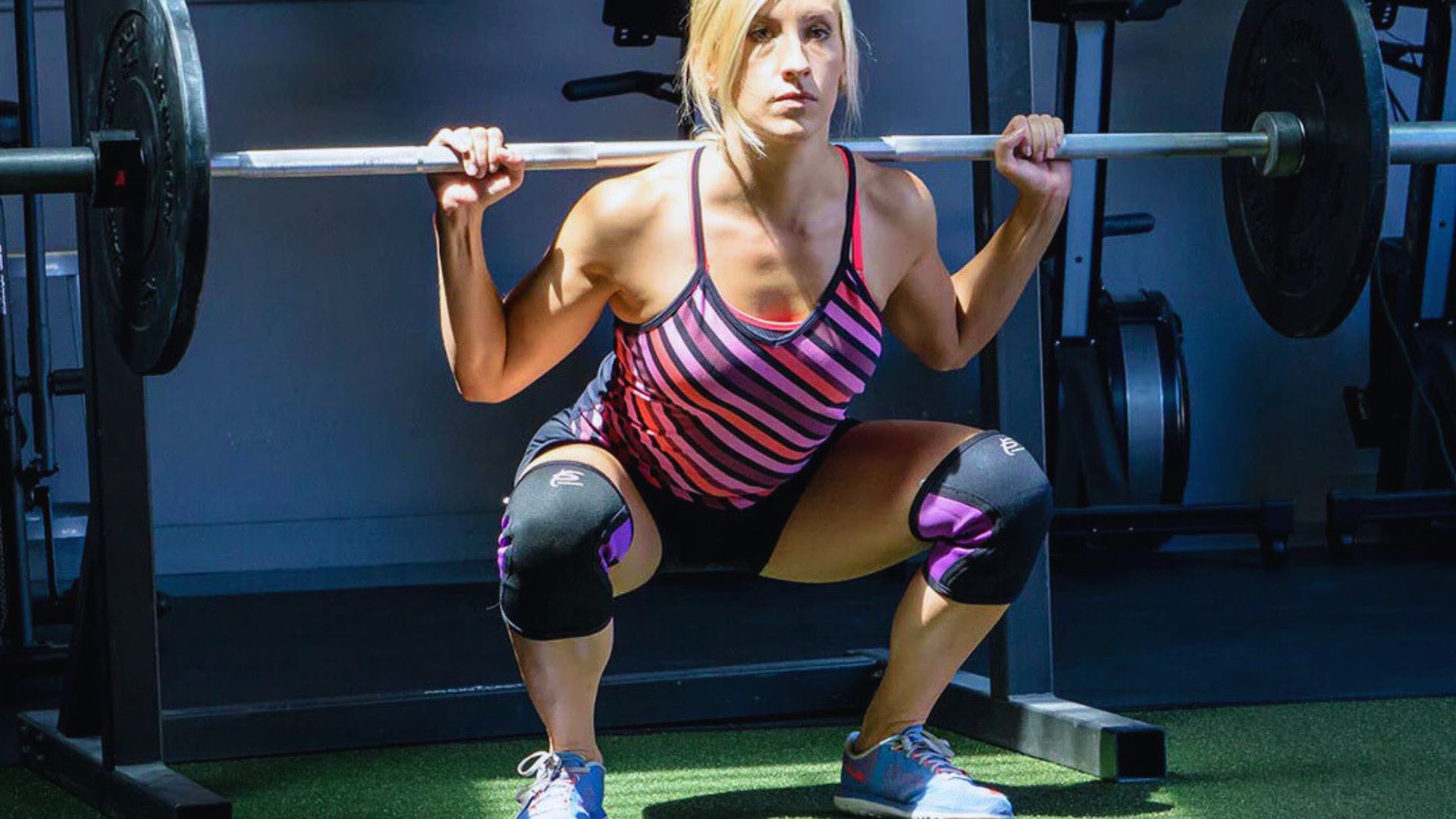 A woman performs a squat with a barbell in a gym. Wearing a striped tank top and knee pads, she appears focused and determined under bright lighting.
