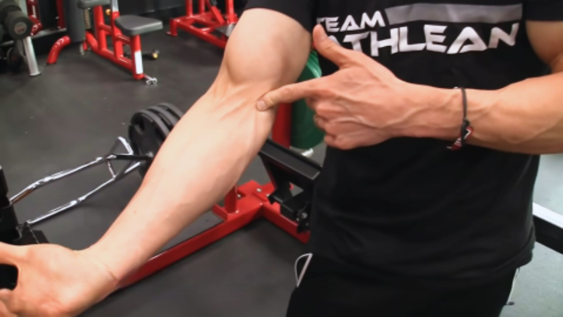 A person in a gym points to their veiny, muscular forearm. They're wearing a black "Team Athlean" shirt. The gym has red and black equipment visible in the background.