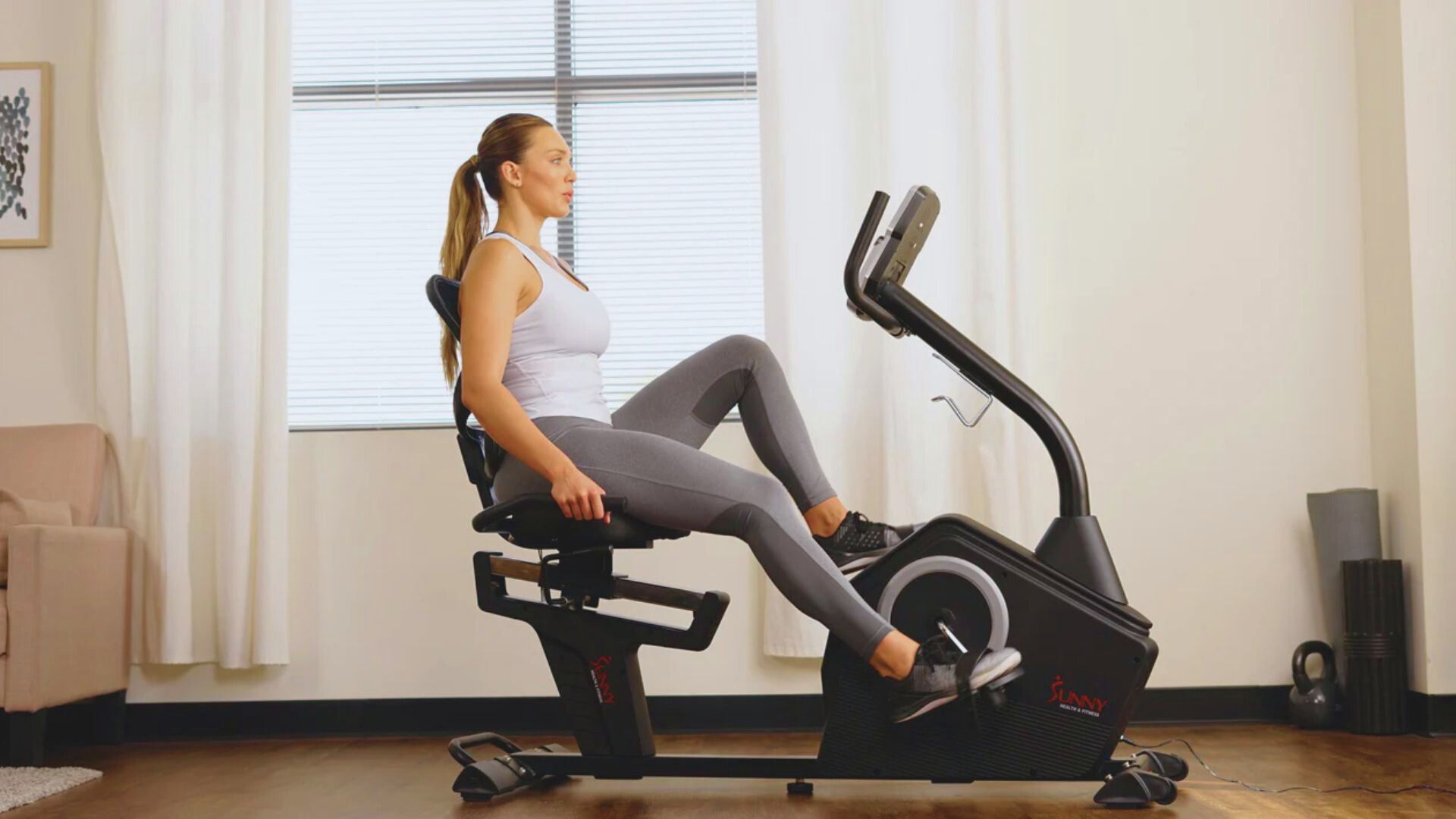 A woman exercises on a recumbent bike in a bright room. She wears a white tank top and gray leggings, concentrating on her workout with a calm expression.