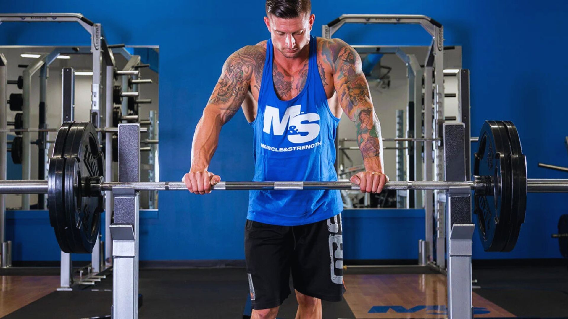 A muscular man in a blue "M&S" tank top grips a barbell on a weightlifting bench in a gym. The room is bright with mirrors and weight machines, conveying focus and strength.