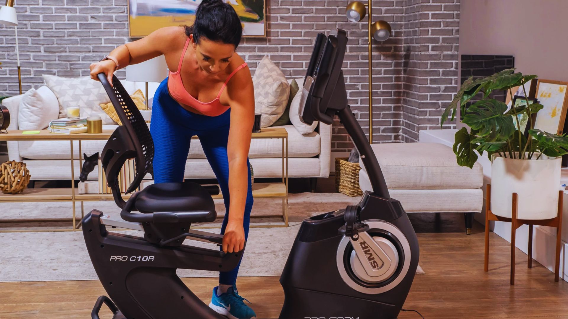 A woman pedals on an exercise bike in a cozy living room, surrounded by plants and natural light.