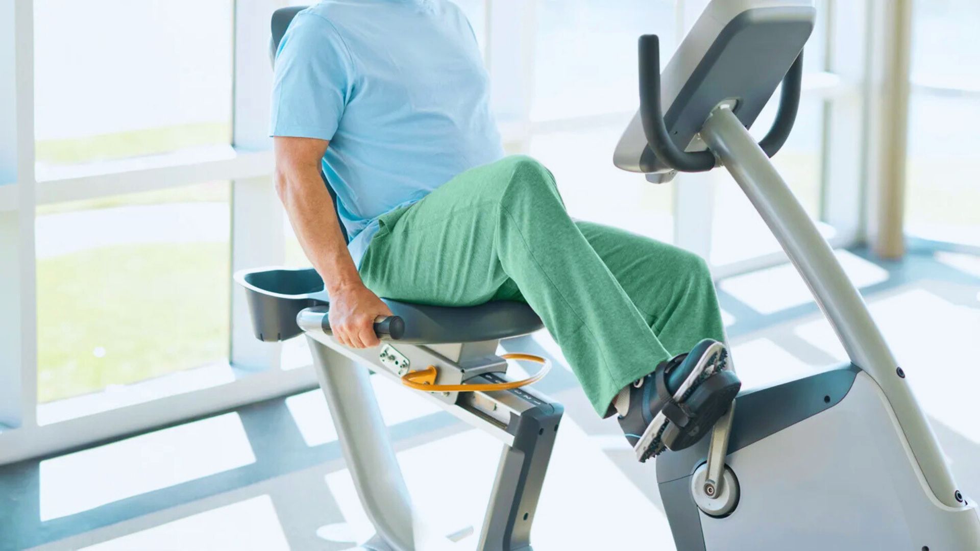 An older adult exercises on a recumbent bike in a bright gym, wearing a blue shirt and green pants. The mood is active and focused.