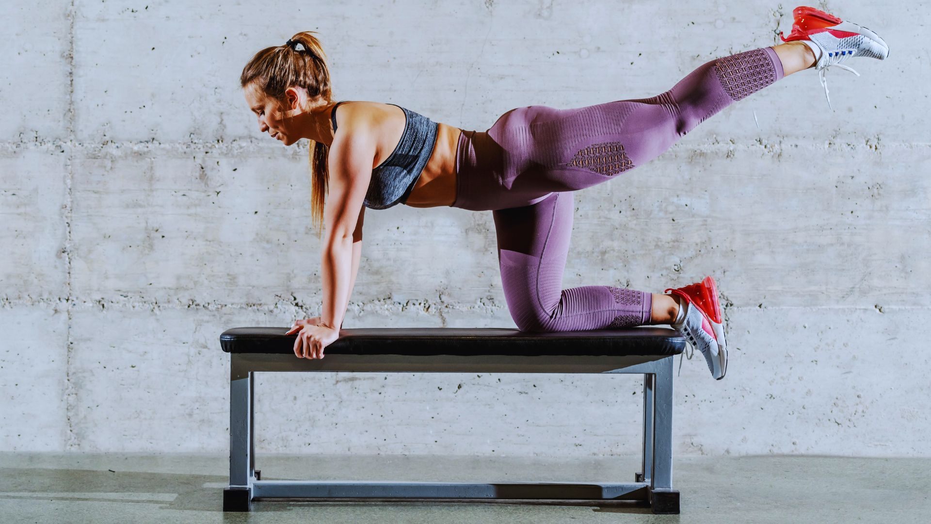 Woman in athletic wear performs a leg lift on a bench against a concrete wall. She exudes strength and balance, embodying focus and determination.