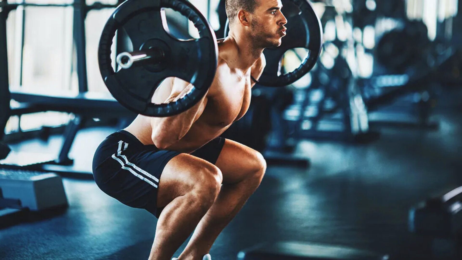 A man in a gym performs a barbell squat. He's focused, wearing athletic shorts. The environment is bright with fitness equipment in the background.