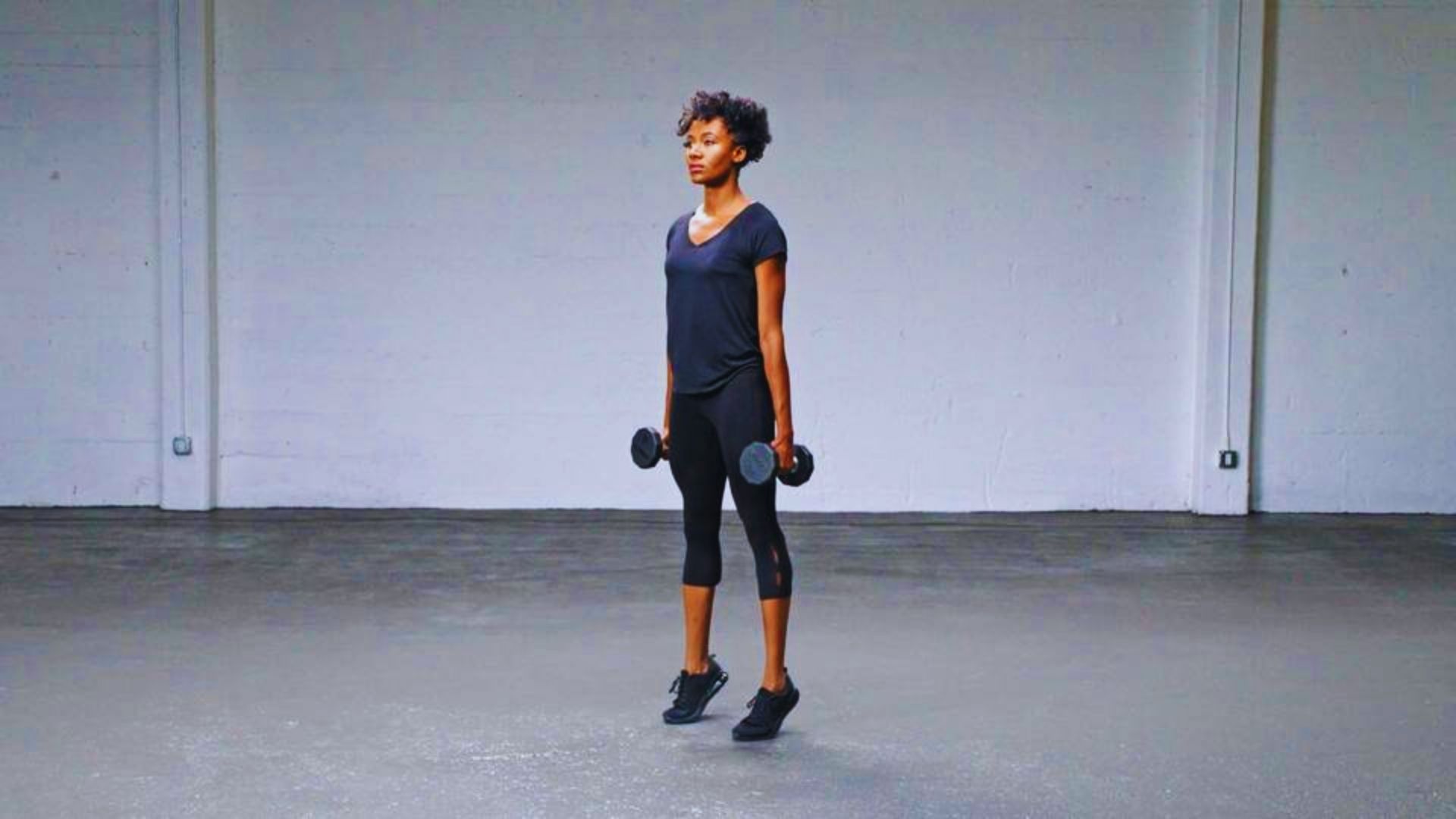 A woman stands in an empty room, holding a dumbbell, ready for a workout.