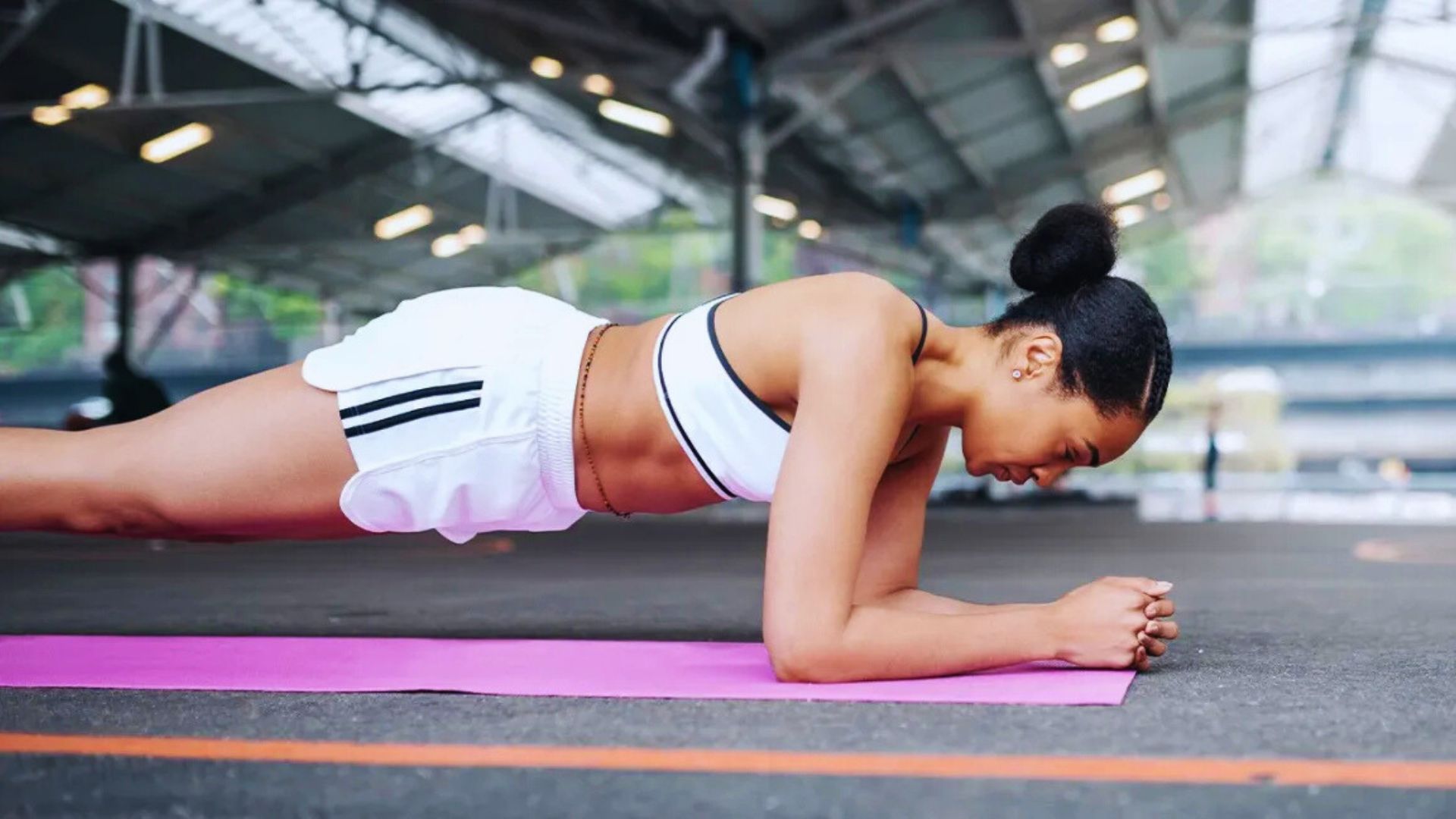 Woman in a white sports outfit holds a plank position on a pink yoga mat under a large, well-lit pavilion. She appears focused and determined.