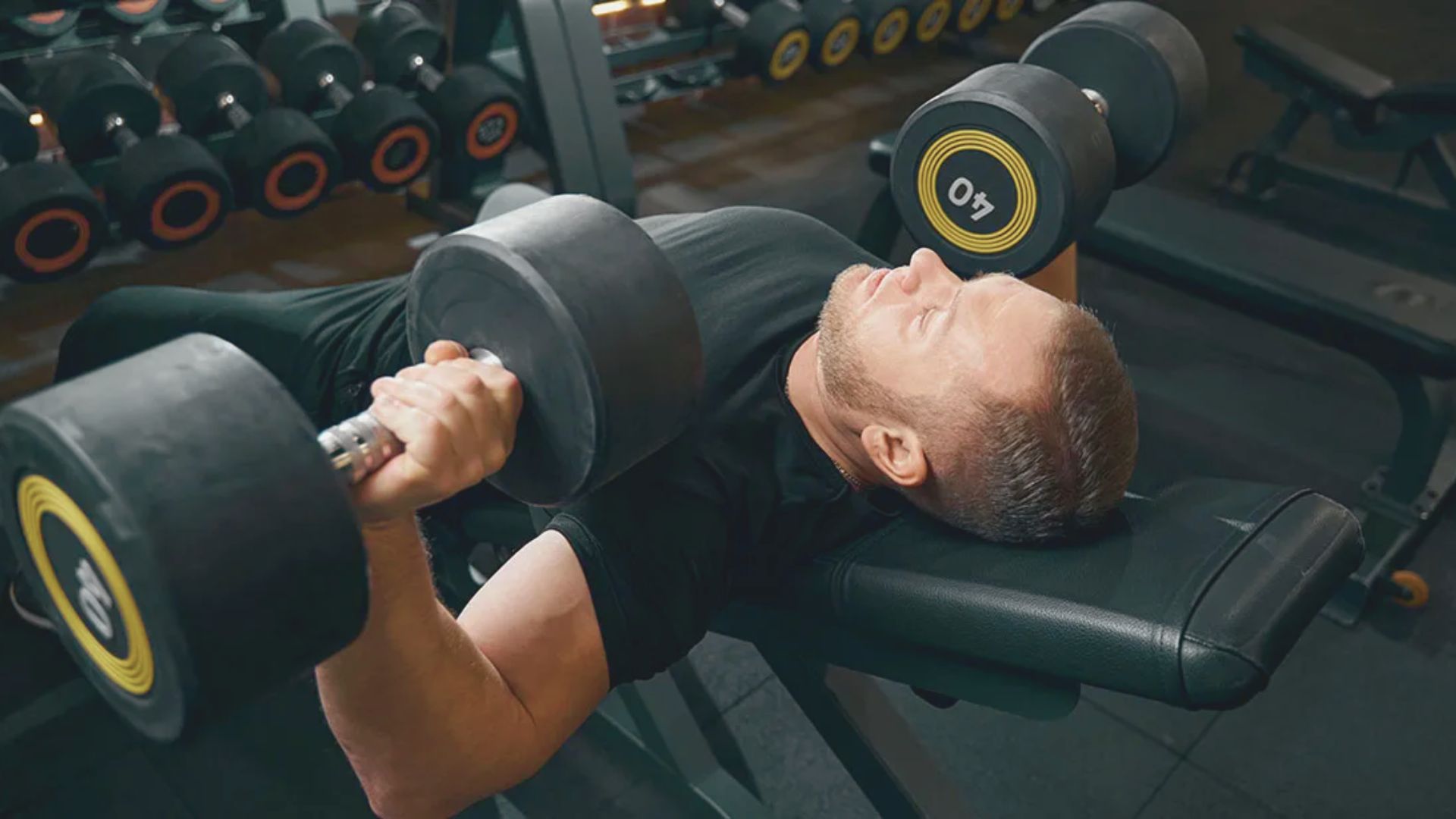 A man lies on a bench, holding a dumbbell in one hand, focused on his workout routine.