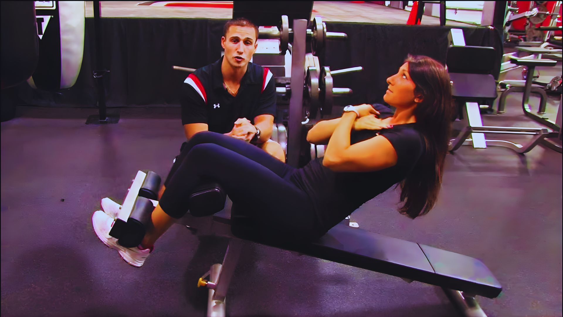 A woman sits on a bench while a man performs a sit-up on the ground nearby, both engaged in fitness activities.