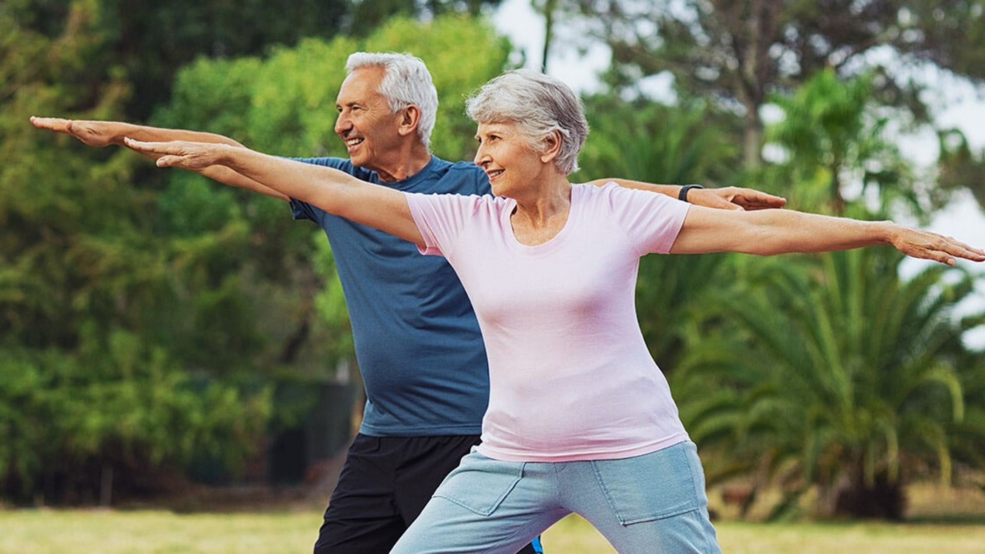 Elderly couple practicing yoga outdoors, smiling in warrior pose. Bright clothing, lush green trees in background, conveying relaxation and vitality.