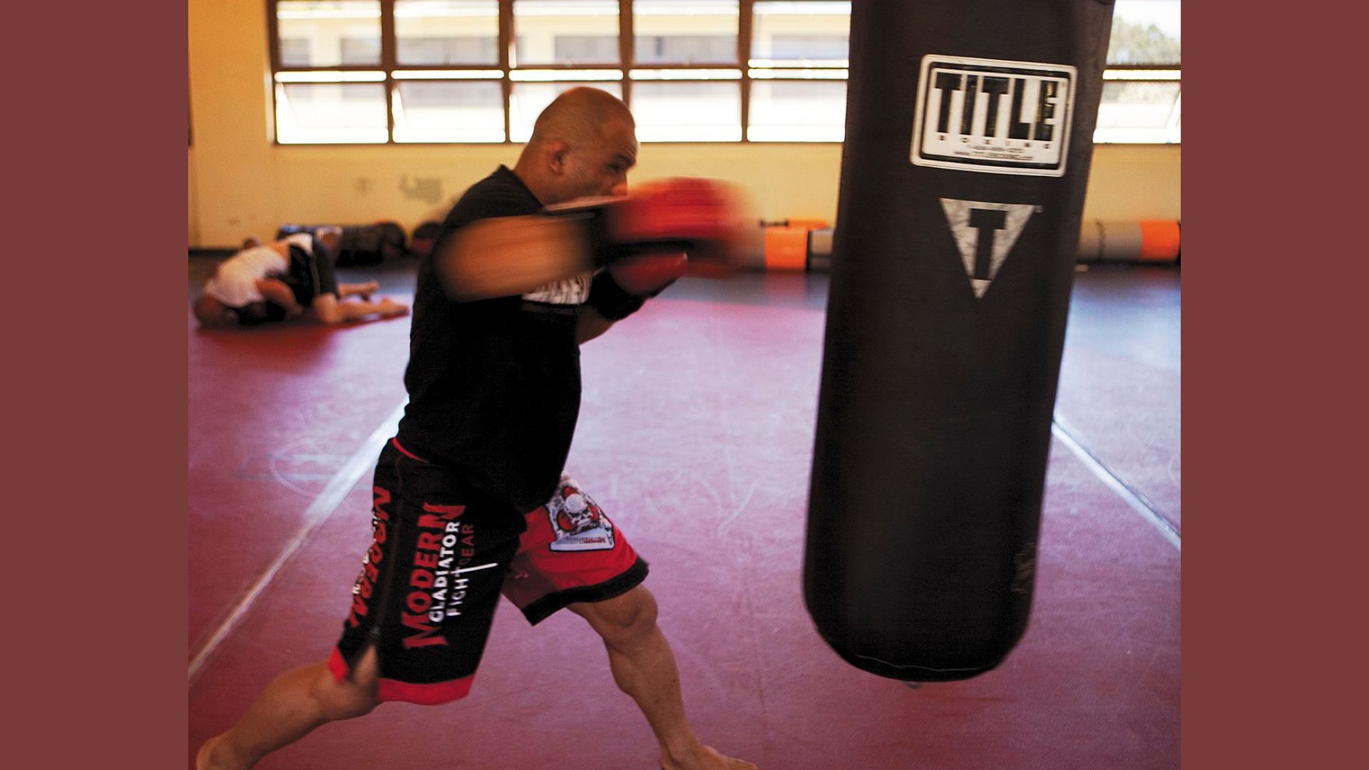 A man in a black shirt and red shorts strikes a punching bag with focused intensity during a workout session.