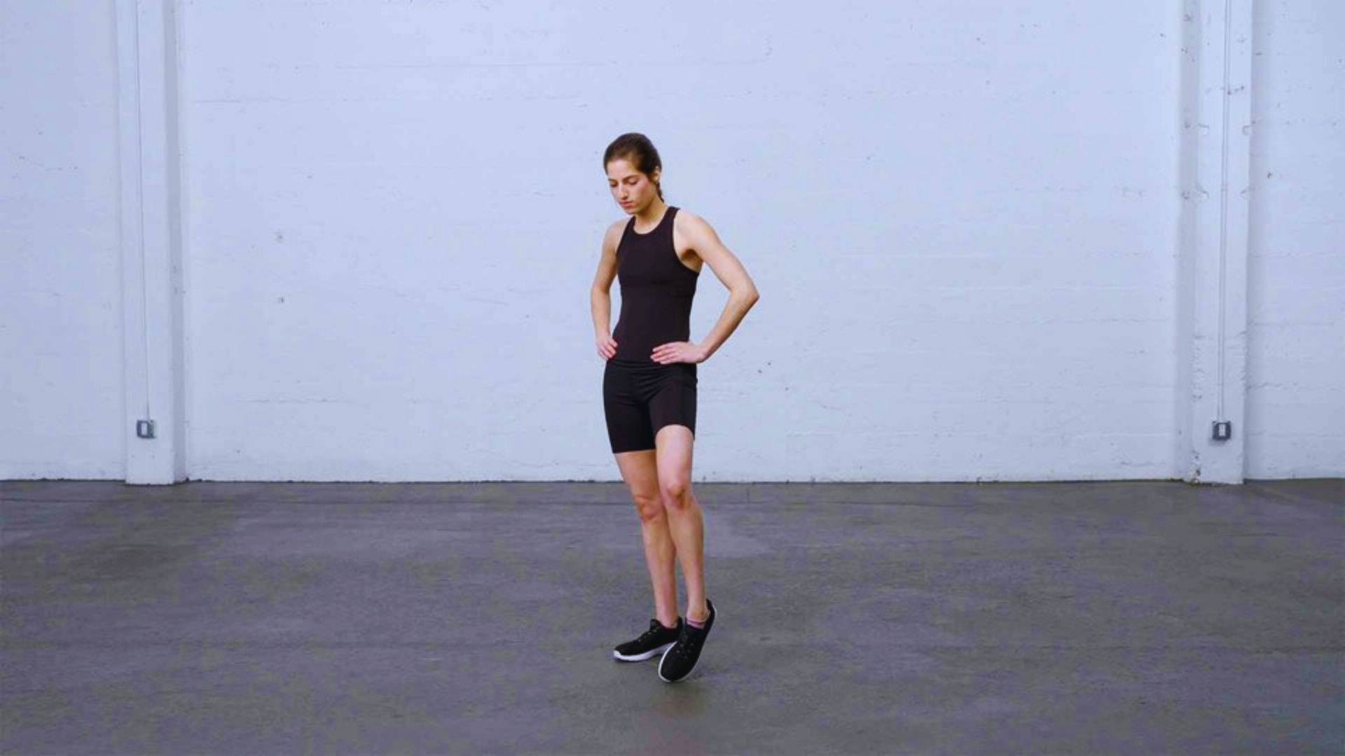 A woman in a black tank top and shorts stands in an empty room, looking around thoughtfully.