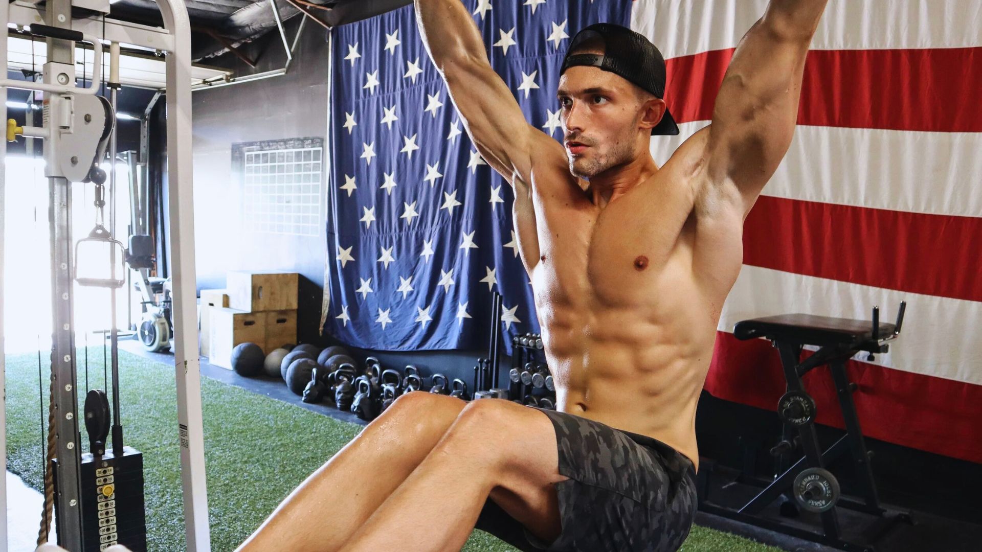 Shirtless man doing pull-ups in a gym, with an American flag backdrop. The setting includes gym equipment and a focused, intense atmosphere.