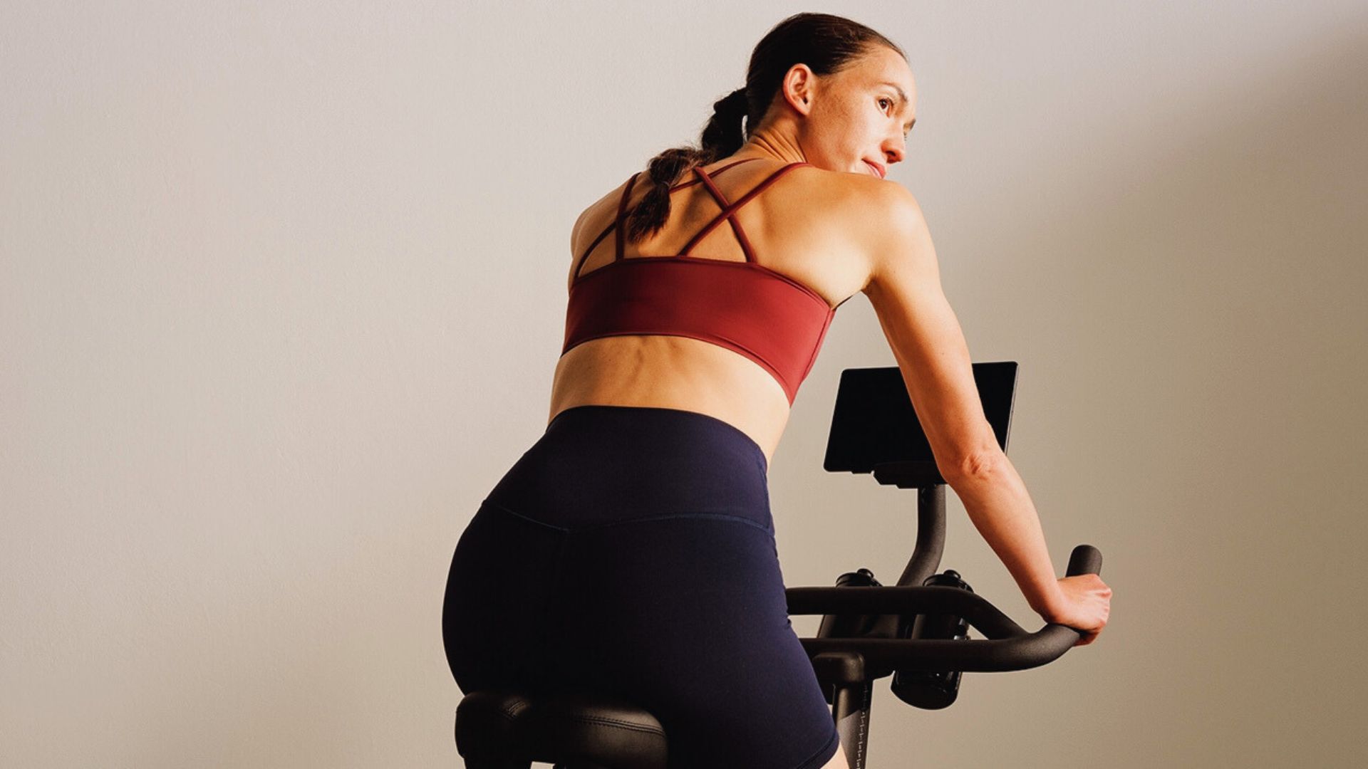 A woman in a red sports bra and black shorts exercises on a stationary bike, looking focused. The scene is minimalist with a neutral background.