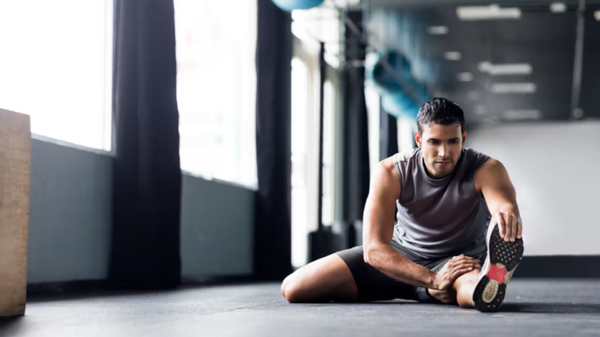 Man in gym attire stretching on the floor, surrounded by exercise equipment. The atmosphere is focused and calm, with natural light streaming in.