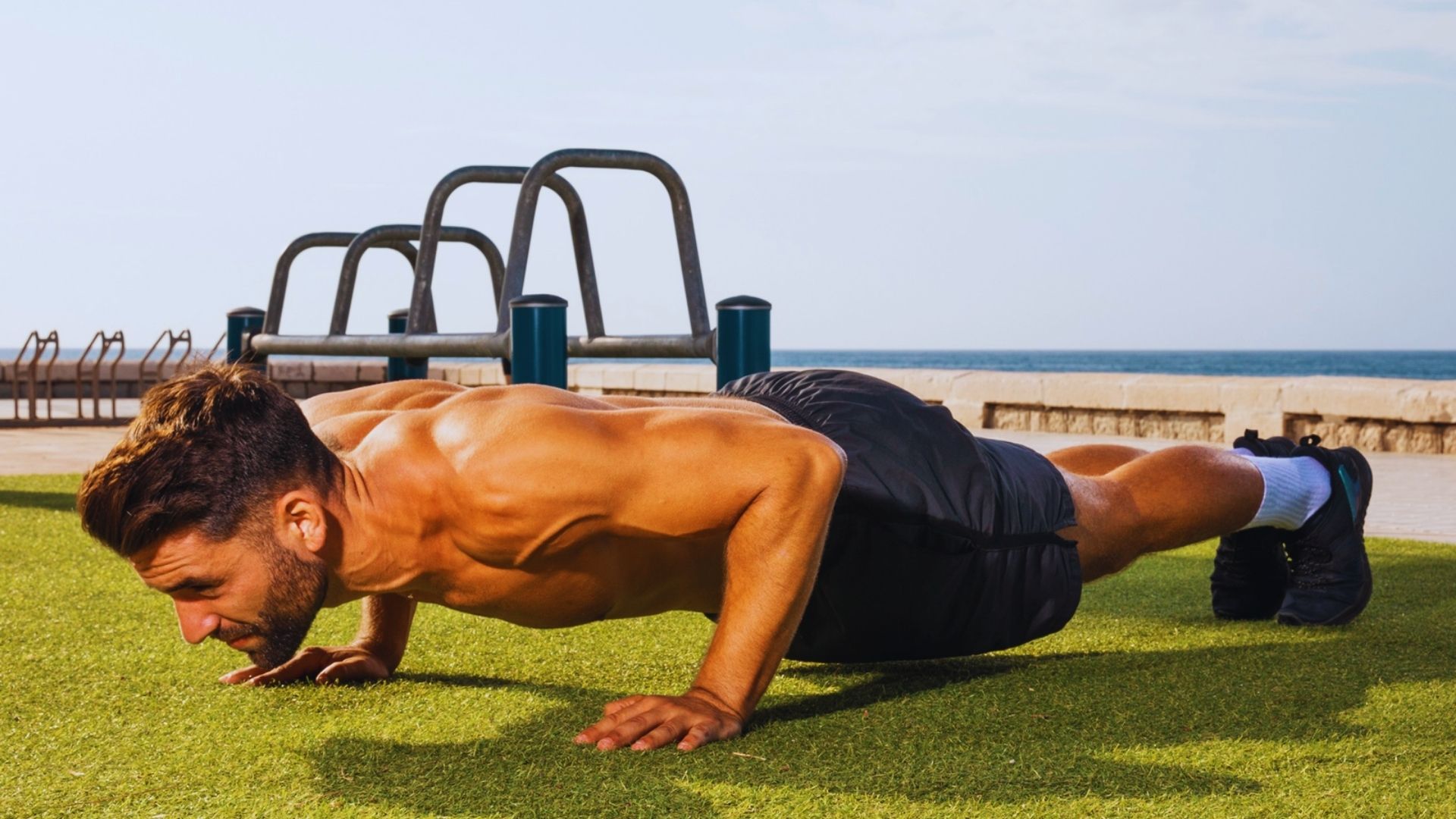 Man performing push-ups on grass with the ocean in the background under a clear blue sky.