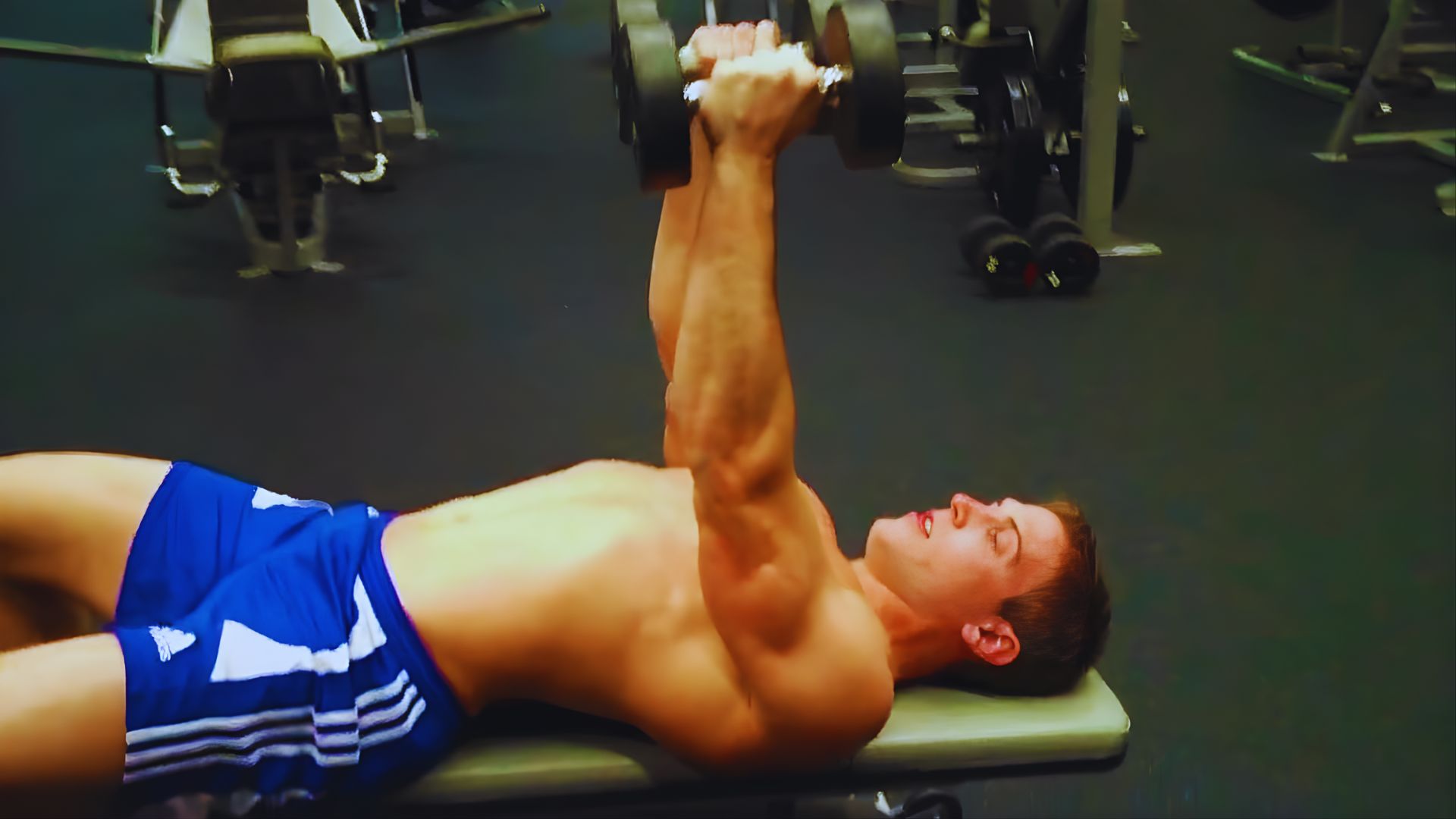 A shirtless man performs a dumbbell exercise, focusing on strength training in a gym setting.